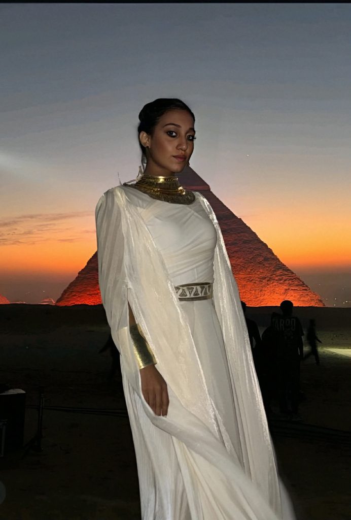 Young woman with long dark hair, brown eyes, a gold necklace, and a white gown, in front of one of Egypt's pyramids at sunset or sunrise. 