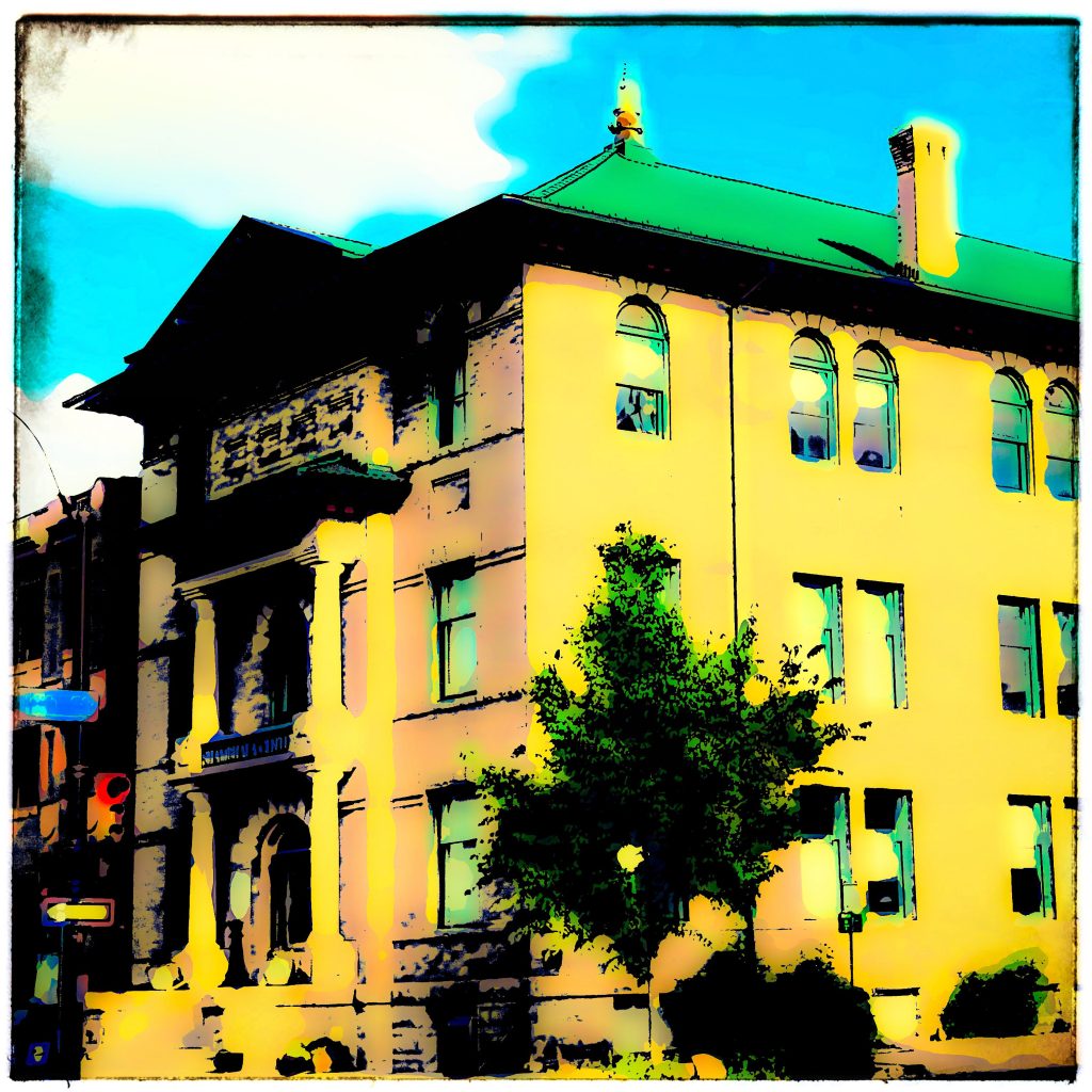 Large three story yellow building with a green roof and tree on a partly cloudy day. 