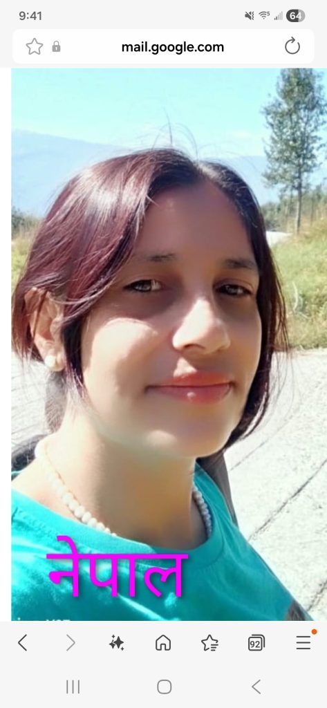 Young South Asian woman with dark hair up in a bun, brown eyes, and a white bead necklace and blue top standing outside near grass. 