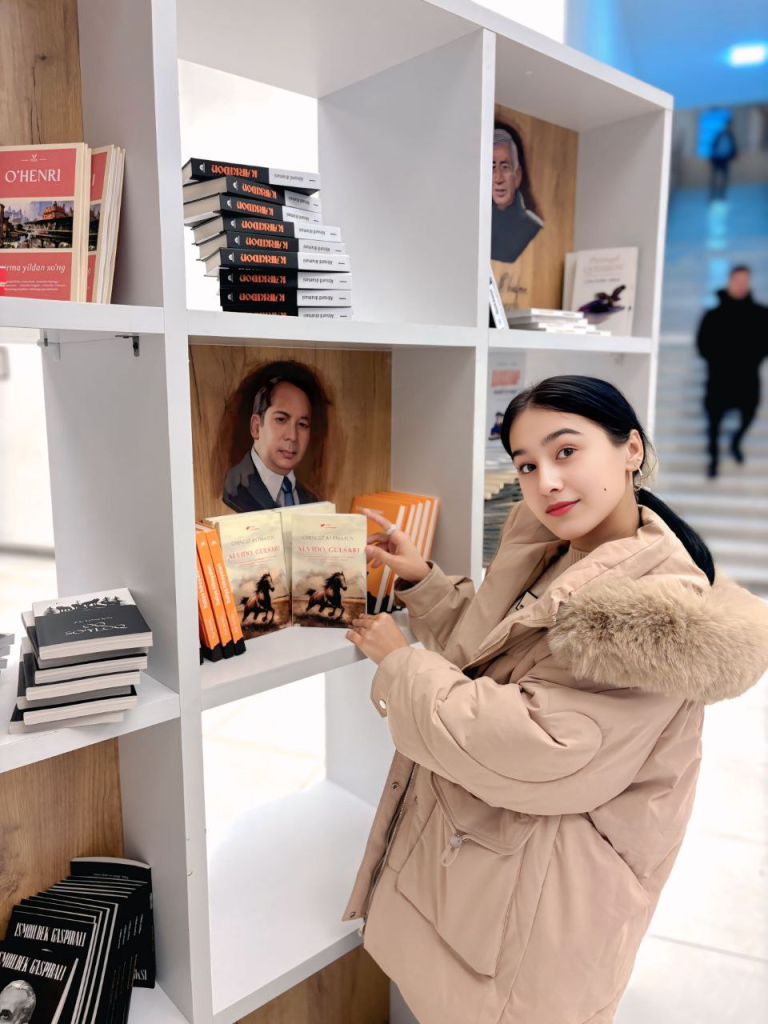 Young Central Asian woman with long dark straight hair up in a bun or ponytail standing near a book on a display case and wearing a pink jacket. 