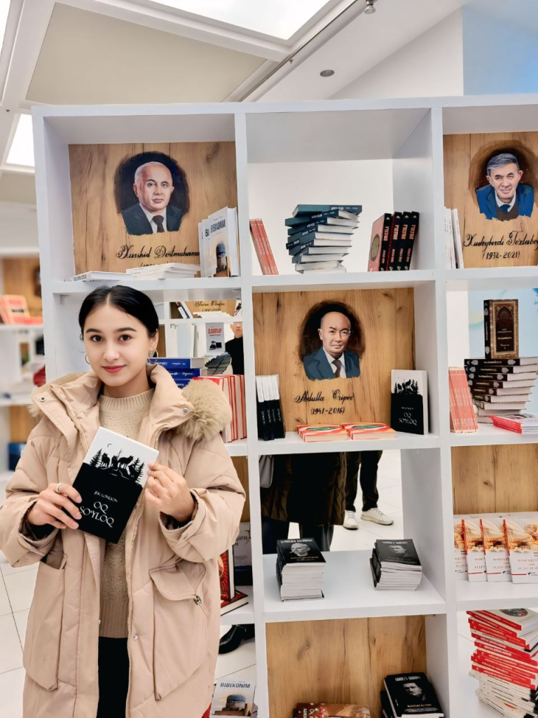 Central Asian woman, young, with dark hair in a bun, hoop earrings, and a tan coat, holding a book.