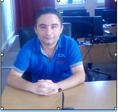 Young middle aged Greek man in a blue polo shirt and short dark hair seated at a wooden desk.