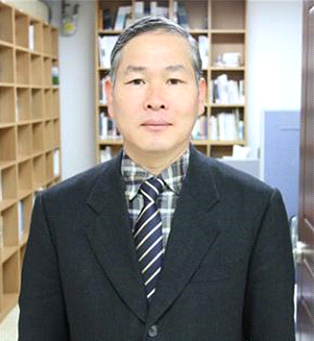 Older middle aged East Asian man standing in a library or office in a dark suit and tie.