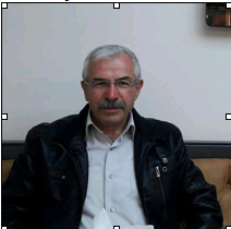 Older South Asian man with gray hair and a gray collared shirt and dark coat.