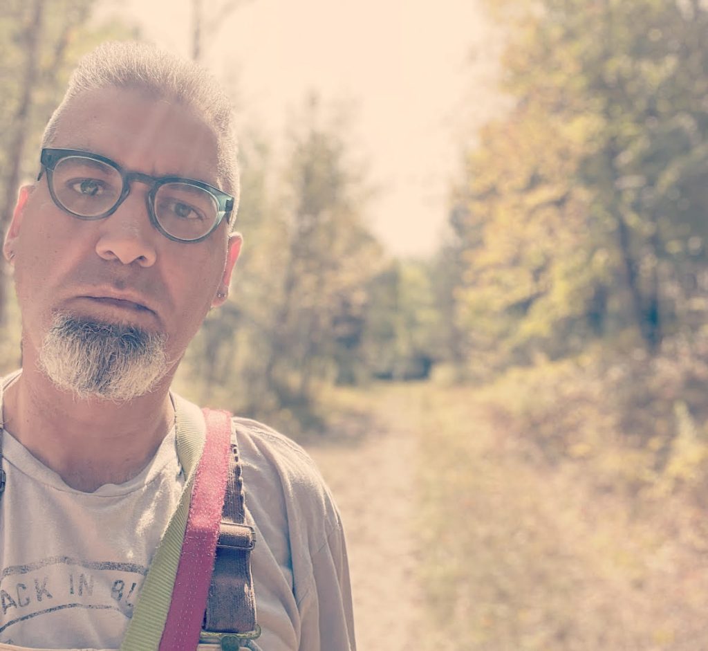 Middle aged white man with reading glasses, a bit of hair and a trimmed beard, in a tee shirt and backpack on a hike out by trees and a trail on a sunny day. 