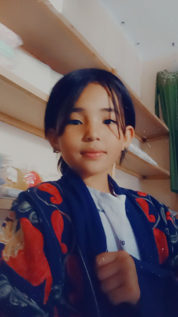 Young Central Asian woman with straight dark hair up in a bun, brown eyes, and a blue and red sweater over a white top. 