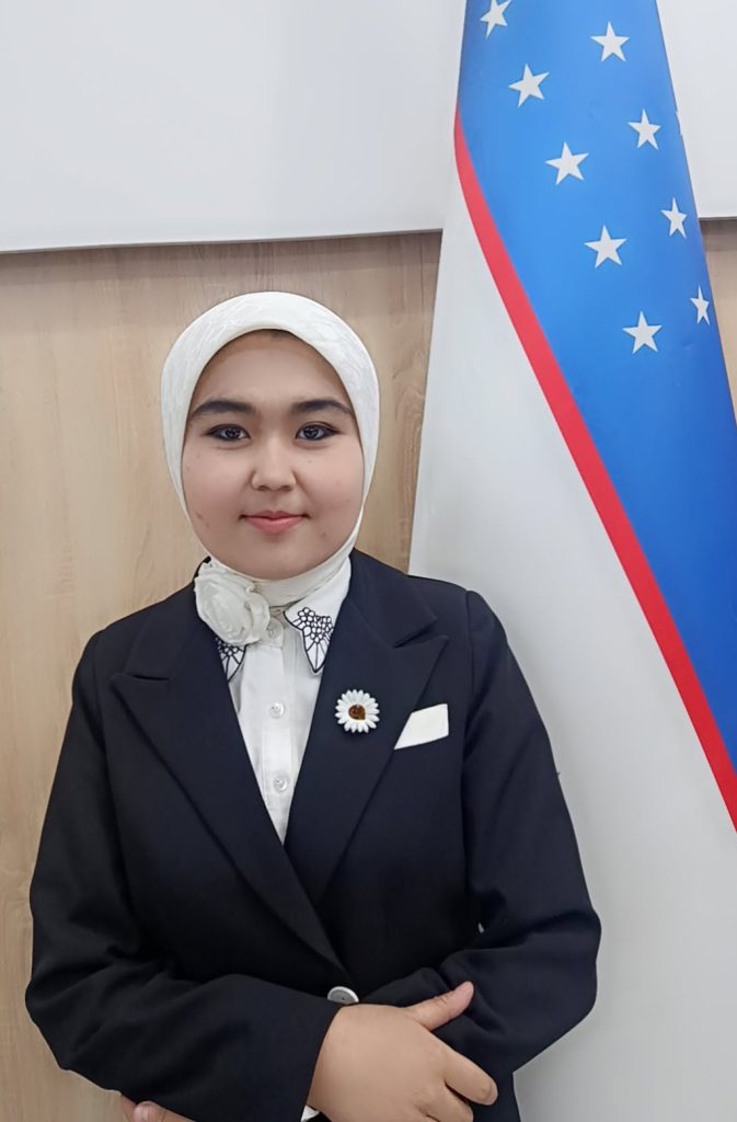 Image of a young Central Asian woman in a black coat, collared blouse, flower pin, and white headscarf standing next to a flag. 