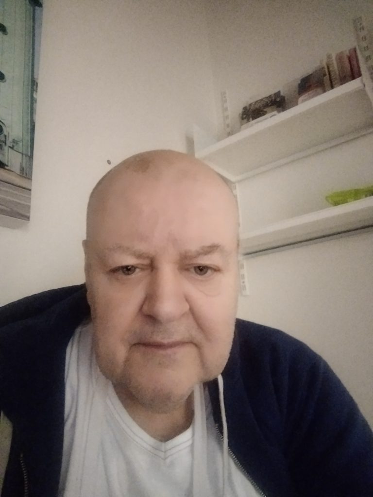 Older white man with not much hair and a white tee shirt and black sweater.