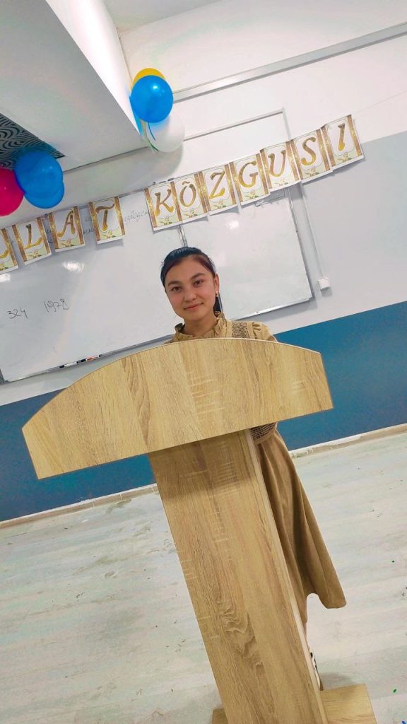 Young Central Asian woman with long dark hair, a long tan dress, standing at a lectern with balloons and signs behind her. 