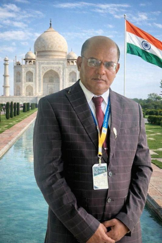 Older middle aged South Asian man in a lanyard and gray suit in front of the Taj Mahal on a sunny day.