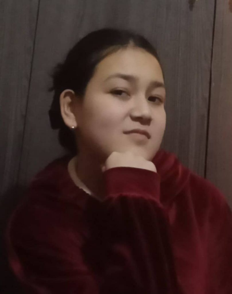 Young Central Asian woman with dark hair up in a bun, brown eyes, and tiny earrings and a burgundy sweater.