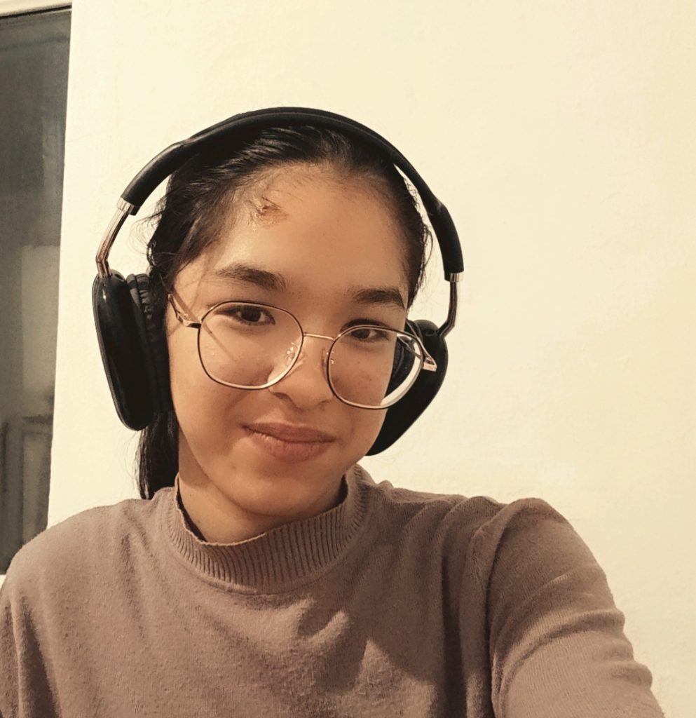 Young Central Asian woman with ear protection, eyeglasses, brown eyes and hair, and a tan sweater.