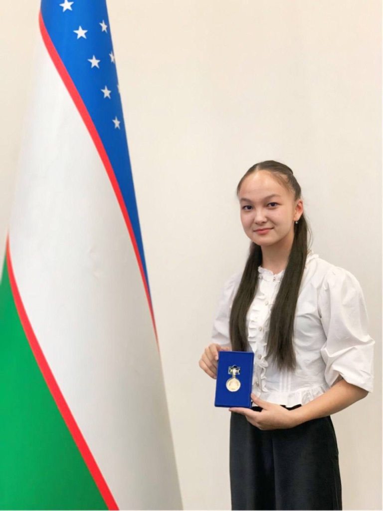 Young Central Asian woman with long dark hair, a white ruffled blouse, and dark black skirt holding up a medal and standing next to a flag. 