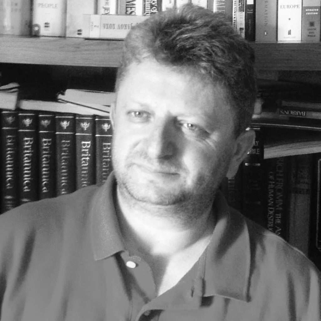 Middle aged white man with short dark hair and a shirt with a collar in front of a shelf of encyclopedias. Black and white image. 