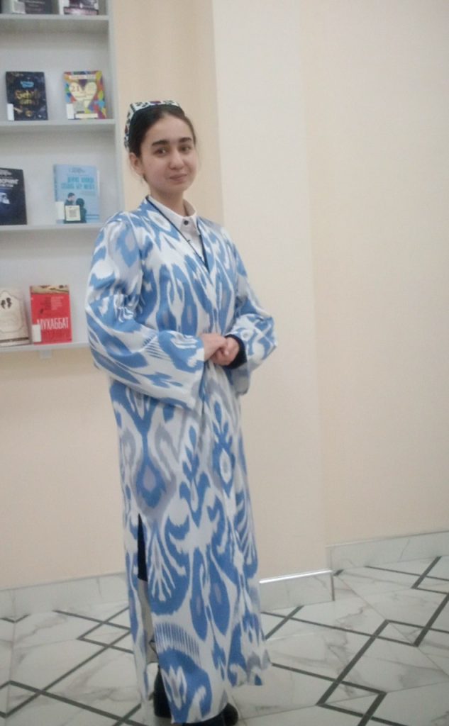 Young Central Asian woman in a patterned white and blue top and embroidered cap standing in front of a bookshelf. 