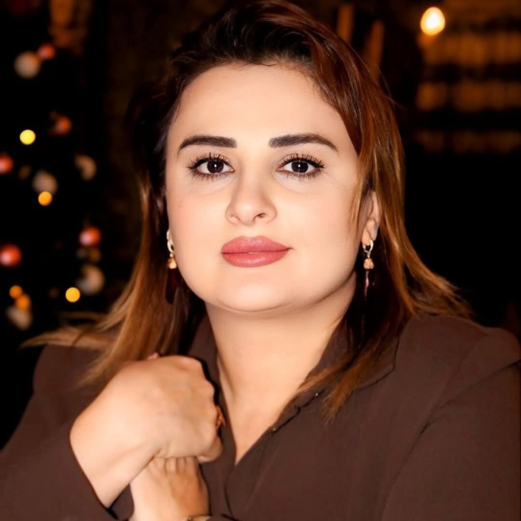 Young light skinned woman with shoulder length brown hair, earrings, and a brown collared sweater. 