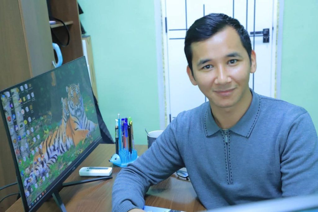 Young Central Asian man with short slick hair, a blue collared top, and a smile, seated by a computer with a tiger  on the screen. 