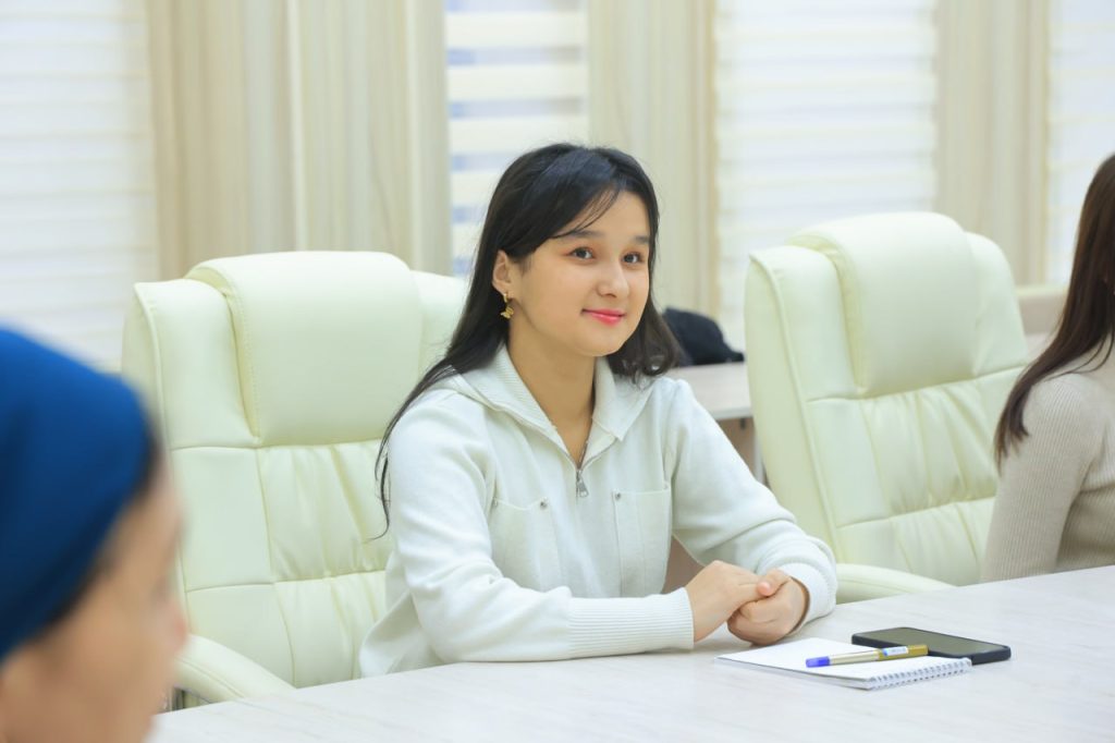 Young Central Asian woman seated at a table with a notebook and pen in front of her. She's in a white collared top with long straight dark hair.