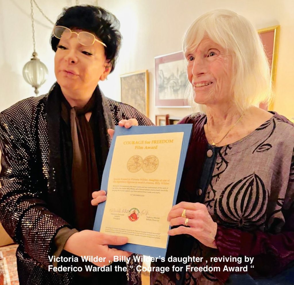 Image of the author with dark hair, a sequined jacket, and reading glasses and a scarf, holding an award and standing next to an old white lady with white hair. 