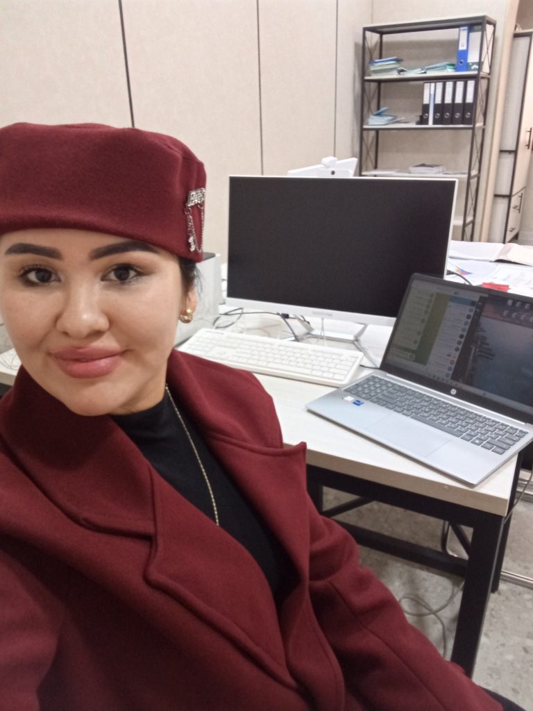 Young Central Asian woman in a burgundy coat and hat seated at a desk with a computer. 