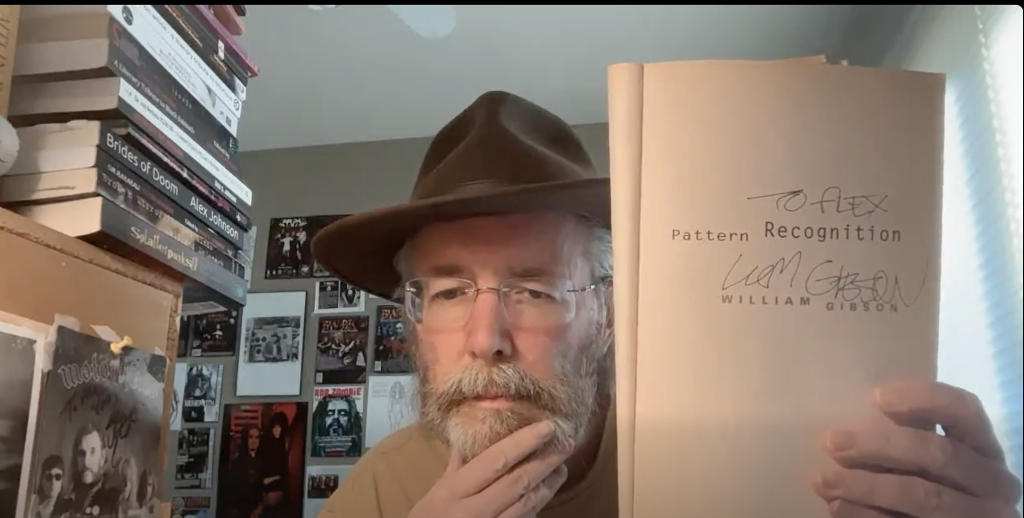 Older white man with reading glasses and a dark colored hat and a trimmed mustache and beard holding up the signed front page of an open book. He's in a room surrounded by books and posters. 