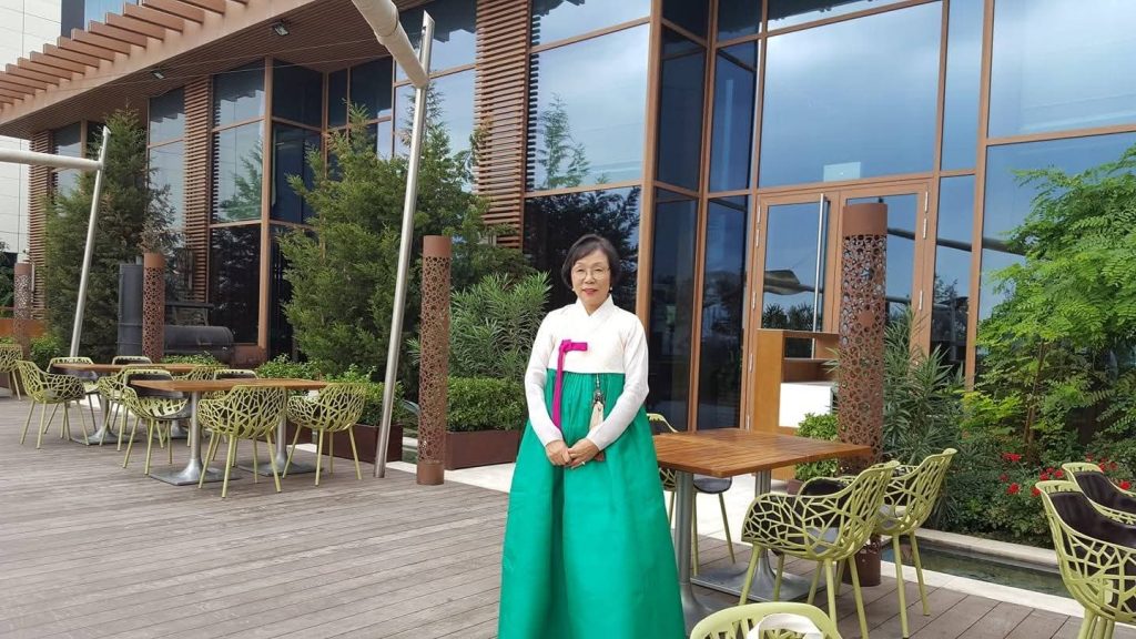 Middle aged Central Asian woman in a long green dress, white top, and black hair standing on a patio in front of a building.