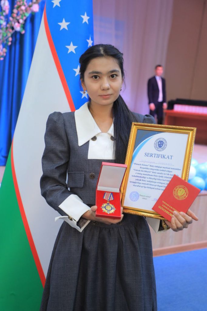 Young Central Asian woman with long black hair and a gray dress with a white collar holding up a certificate standing in front of a flag.
