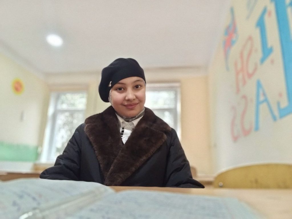 Young Central Asian woman with a black cap, dark fur-lined coat, seated at a desk.