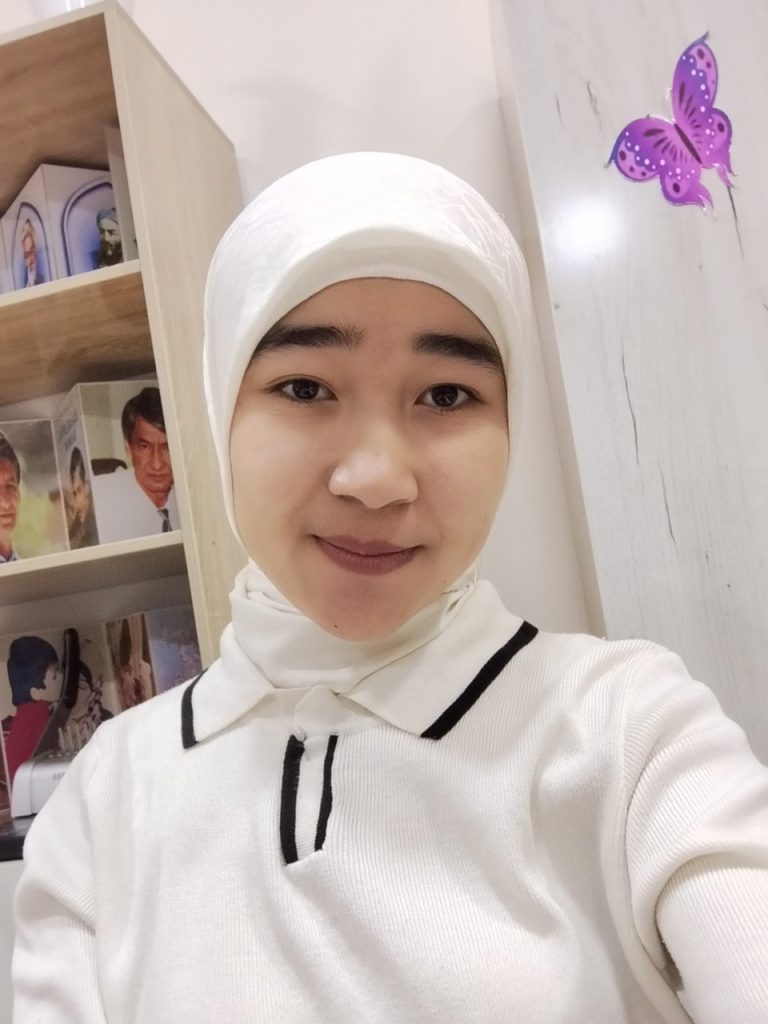Young Central Asian woman in a white headscarf and white outfit with black trim standing in front of a bookshelf.
