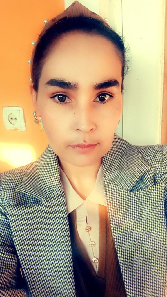 Young Central Asian woman with a tan headscarf, brown eyes, earrings, and a black and white checkered coat.