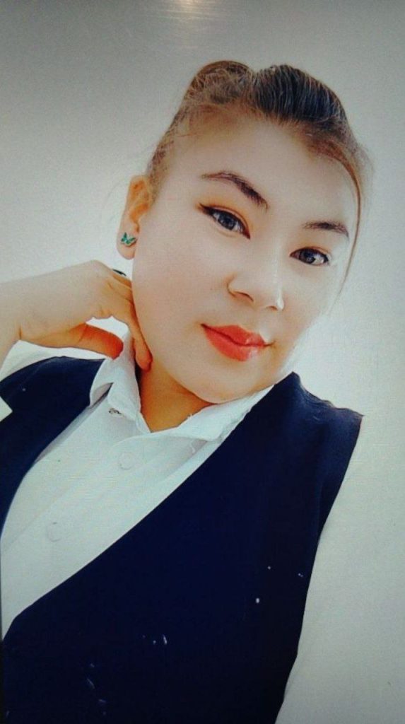 Young Central Asian woman with straight dark hair up in a bun. Small earrings, white collared top, black vest.