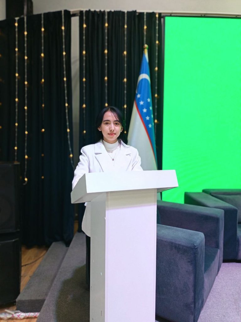 Central Asian woman with short dark hair and a white top and coat standing at a podium next to a flag and some gray chairs and a green screen. 