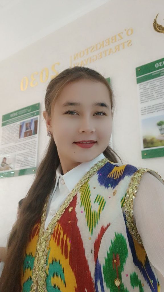 Young Central Asian woman with long dark hair up in pigtails. She's got small earrings, a white collared shirt, and a multicolored vest.