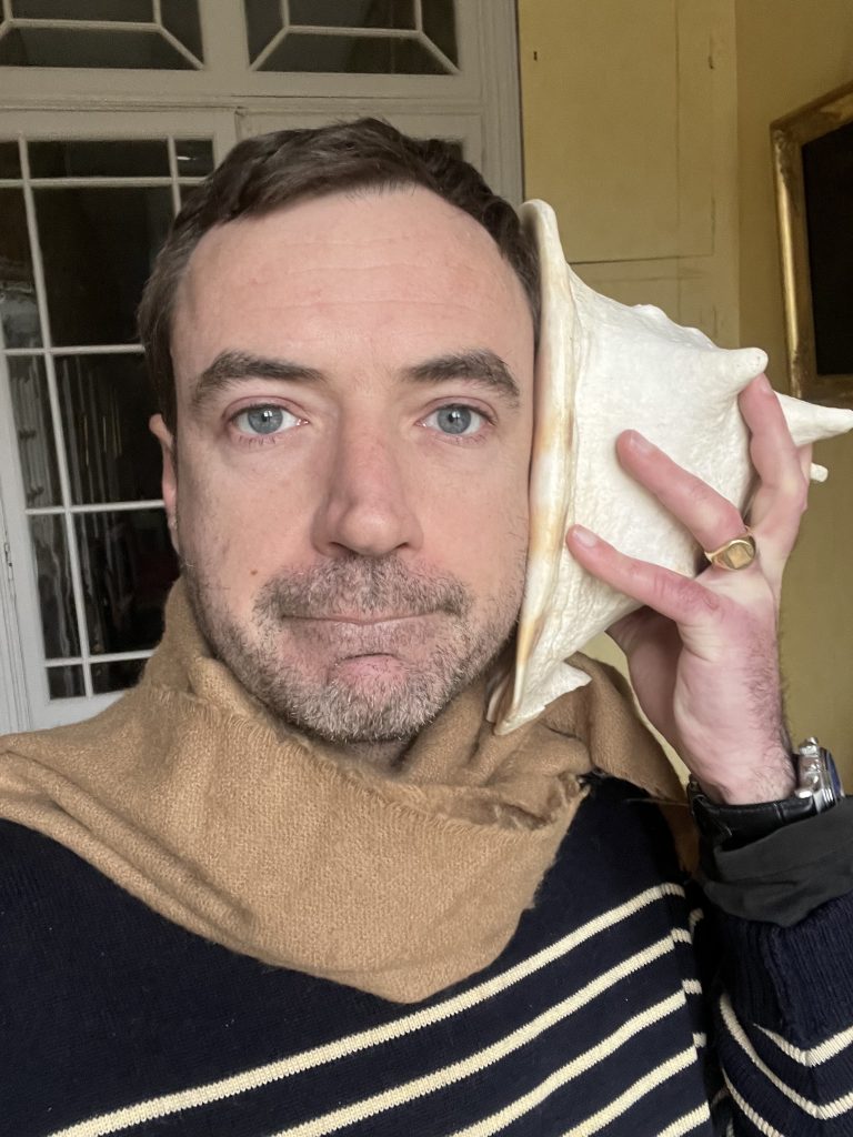 Young white French man with short brown hair, stubble of a beard and mustache, and a brown scarf, holding a giant seashell up to his ear.