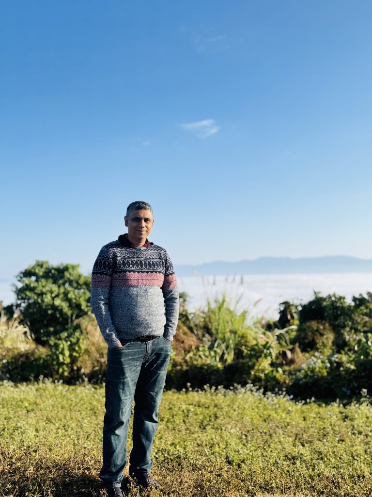 Middle aged Middle Eastern man in a gray and red and black sweater and blue jeans photographed outside on a sunny day in a lawn near some bushes with water and hills in the distance. 