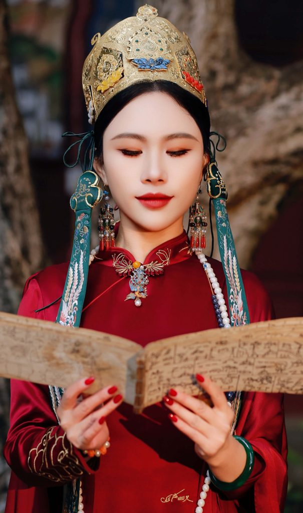 Young Central Asian woman with green headbands and earrings in a red silk gown reading an open book with script text.