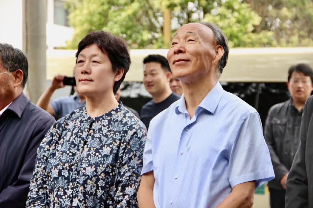 Older East Asian couple standing together. Woman has a flowered top and the man has a blue collared shirt.
