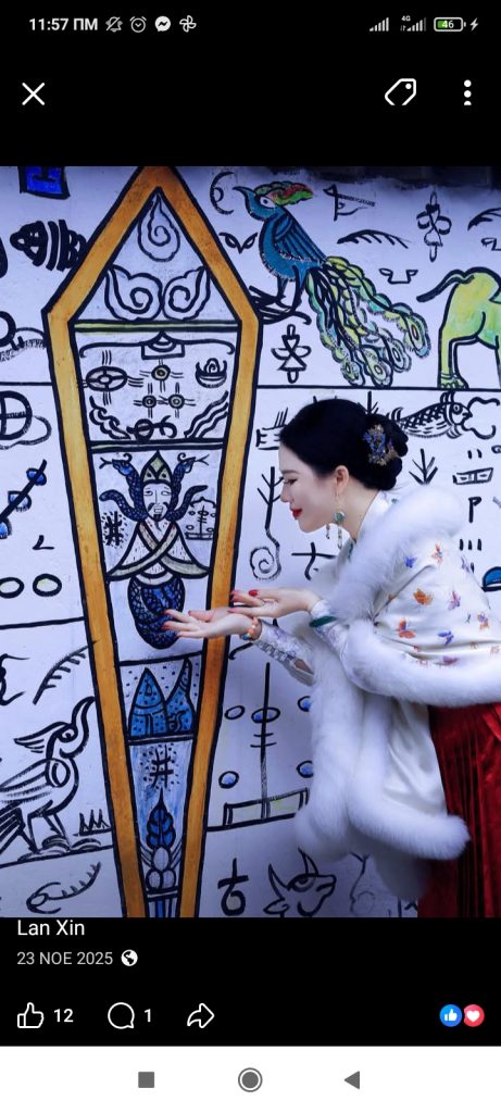Young East Asian woman in a long white dress with butterflies and her hair up in a bun standing in front of a large mural full of line drawings.