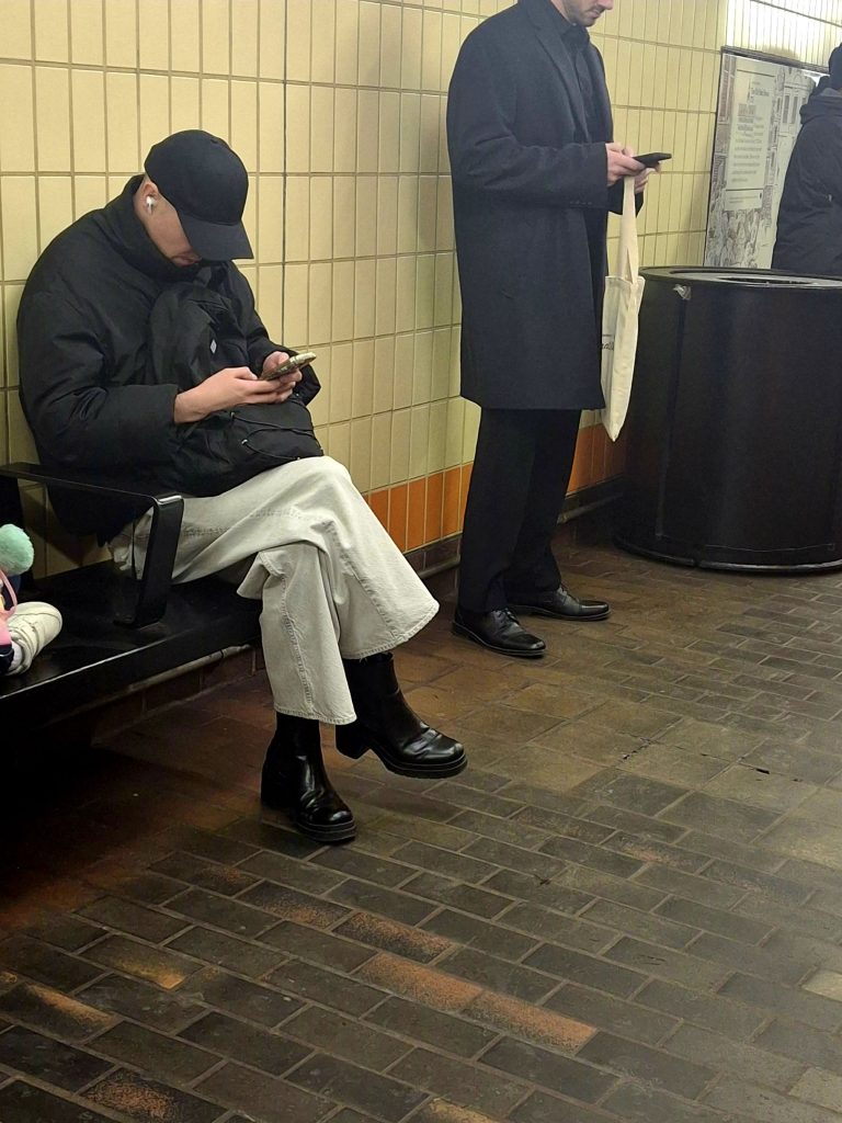 Middle aged person with a baseball cap on and a black jacket and tan jeans and black boots checking their phone in a subway station. Another person of indeterminate gender next to them also checking their phone.