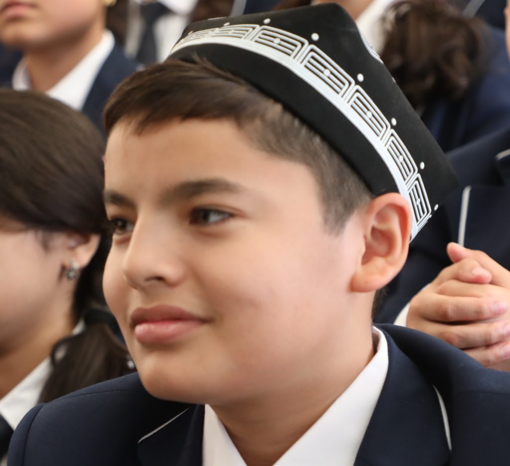Young Central Asian man with a white and black cap, brown hair and eyes, and black coat over a white shirt. 