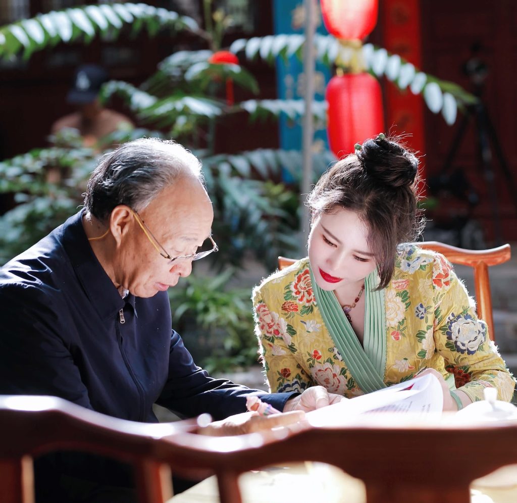 Young East Asian woman with her dark hair up in a bun and a yellow flowered dress seated next to an older East Asian man in reading glasses and a dark coat reading together under the trees.