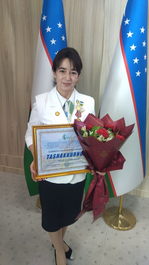 Middle aged Central Asian woman with short dark hair, in a white suit with medals, holding a certificate and bouquet of flowers standing at a podium with flags. 