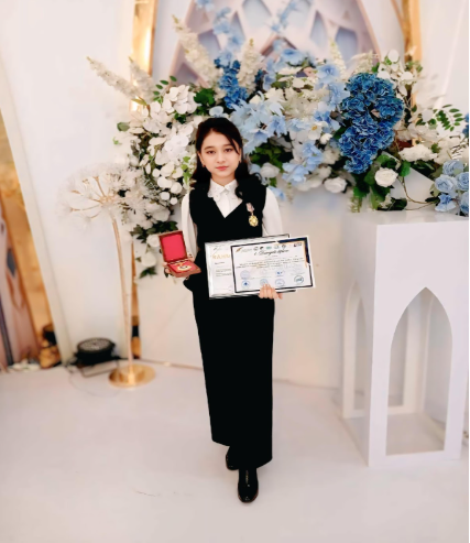 Young Central Asian woman with straight dark hair and a black suit holding a certificate and medal standing indoors in front of many blue and white flowers.