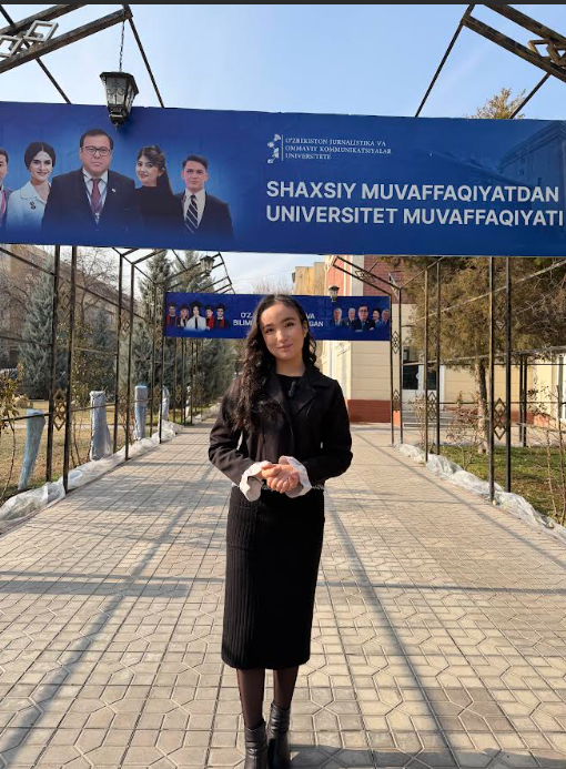 Young Central Asian woman with long dark curly hair in a long black dress and black coat standing on a concrete pathway on her college campus.