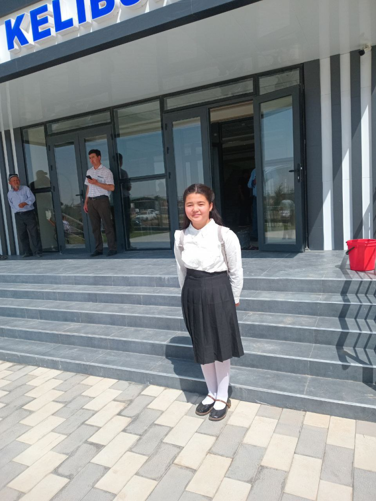 Young Central Asian woman with a white collared shirt, black skirt, and black shoes standing in front of concrete steps leading up into a building. 