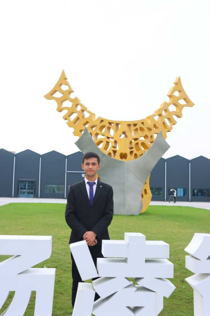 Central Asian man in a black suit and blue tie standing on a green lawn next to some concrete sculptures. 