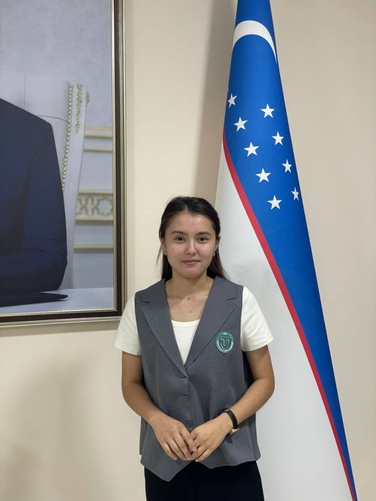 Young Central Asian woman in a gray vest over a white tee shirt standing next to a flag. 