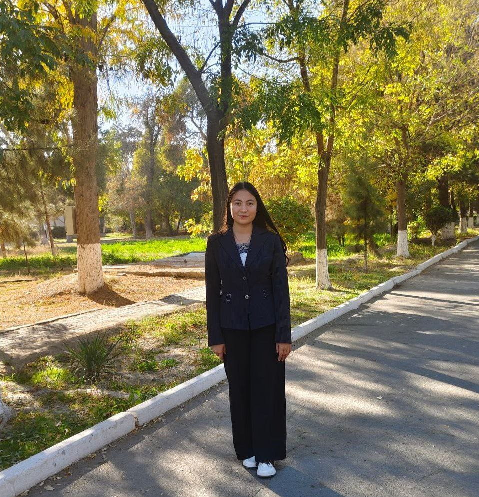 Young Central Asian woman in a black coat and black pants standing on a road near a park with lots of trees on a sunny day.