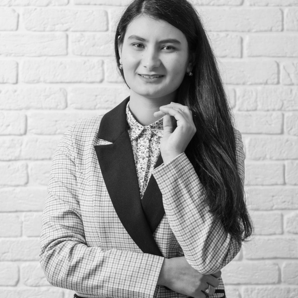 Black and white image of a young Central Asian woman with a collared light colored striped jacket over a flowered collared top. She's got long straight dark hair. 