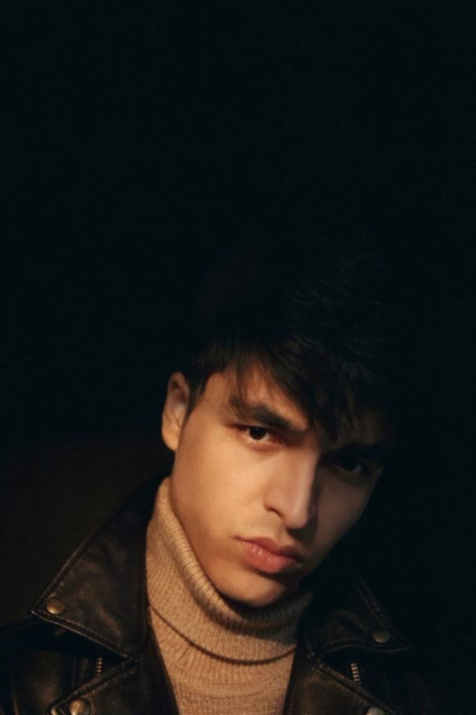 Young Central Asian man in a dark leather jacket, brown eyes and hair, and a tan sweater. 